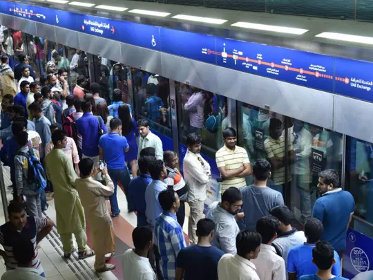 A crowded Dubai Metro platform filled with commuters standing close together while waiting for the train. The metro doors are open, and people are packed near the edge of the platform. Bright overhead lights and a blue station signboard with route information are visible above the train. The scene shows a typical busy workday rush with heavy footfall.