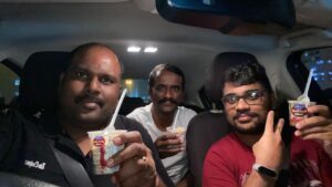 Three friends sitting inside a parked car in Dubai at night, holding small pudding cups and smiling at the camera. The person on the right is me, wearing glasses and a red t-shirt. The car interior lights are on, and the city lights are visible through the windows.