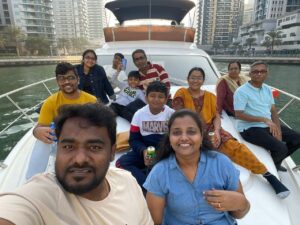 A group of people sitting together on the front deck of a boat in Dubai Marina. High-rise buildings stand in the background. Everyone looks relaxed and smiling, holding drinks and enjoying the boat ride.