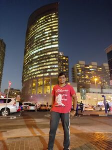 Me standing on a Dubai street at night with tall, brightly lit buildings behind me. I’m wearing a red T-shirt and glasses, and the city lights make everything look lively.